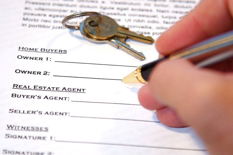 Image of a contract of a home sale, with a hand holding a pen ready to sign the document, and house keys place on top of the paperwork.